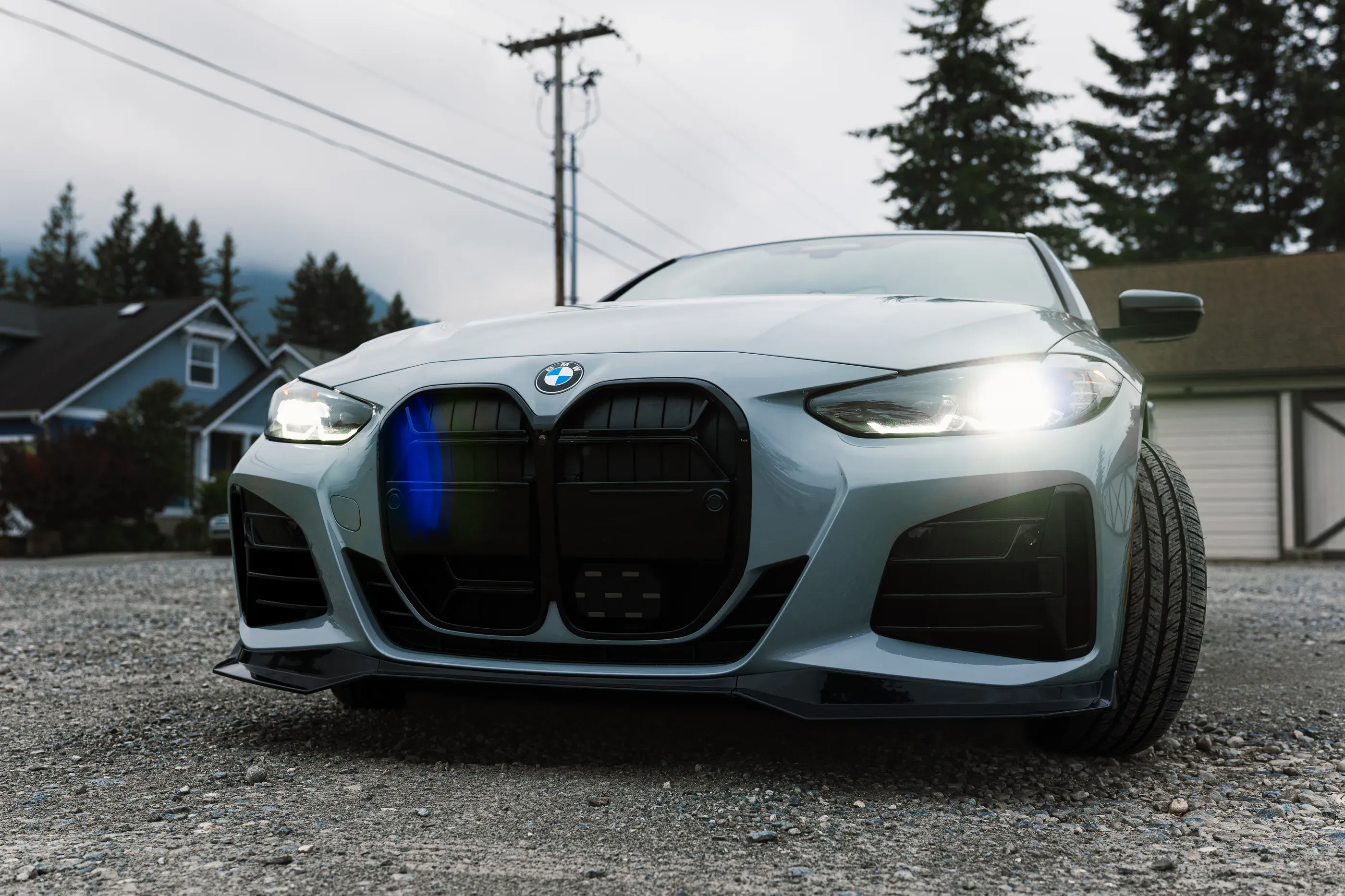 Front view of a grey BMW M440i with headlights on, on a gravel driveway with evergreens behind.