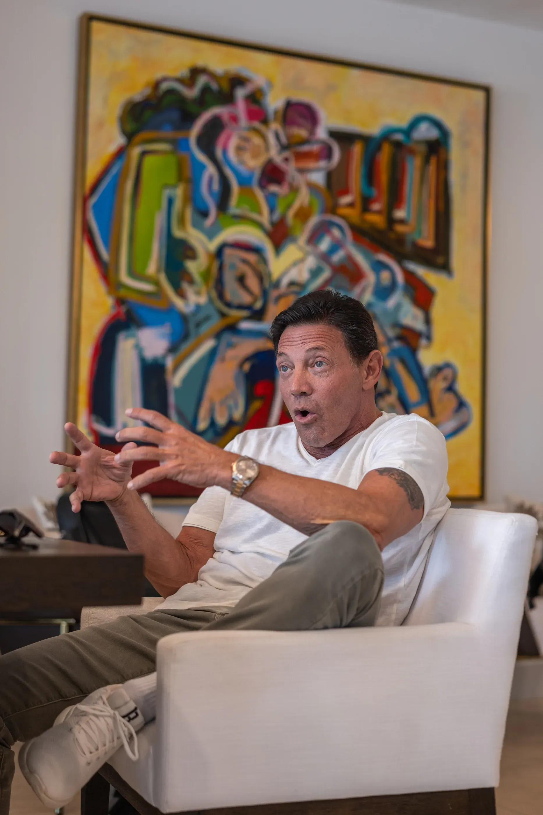 Jordan Belfort talking from an armchair in front of a large abstract painting.