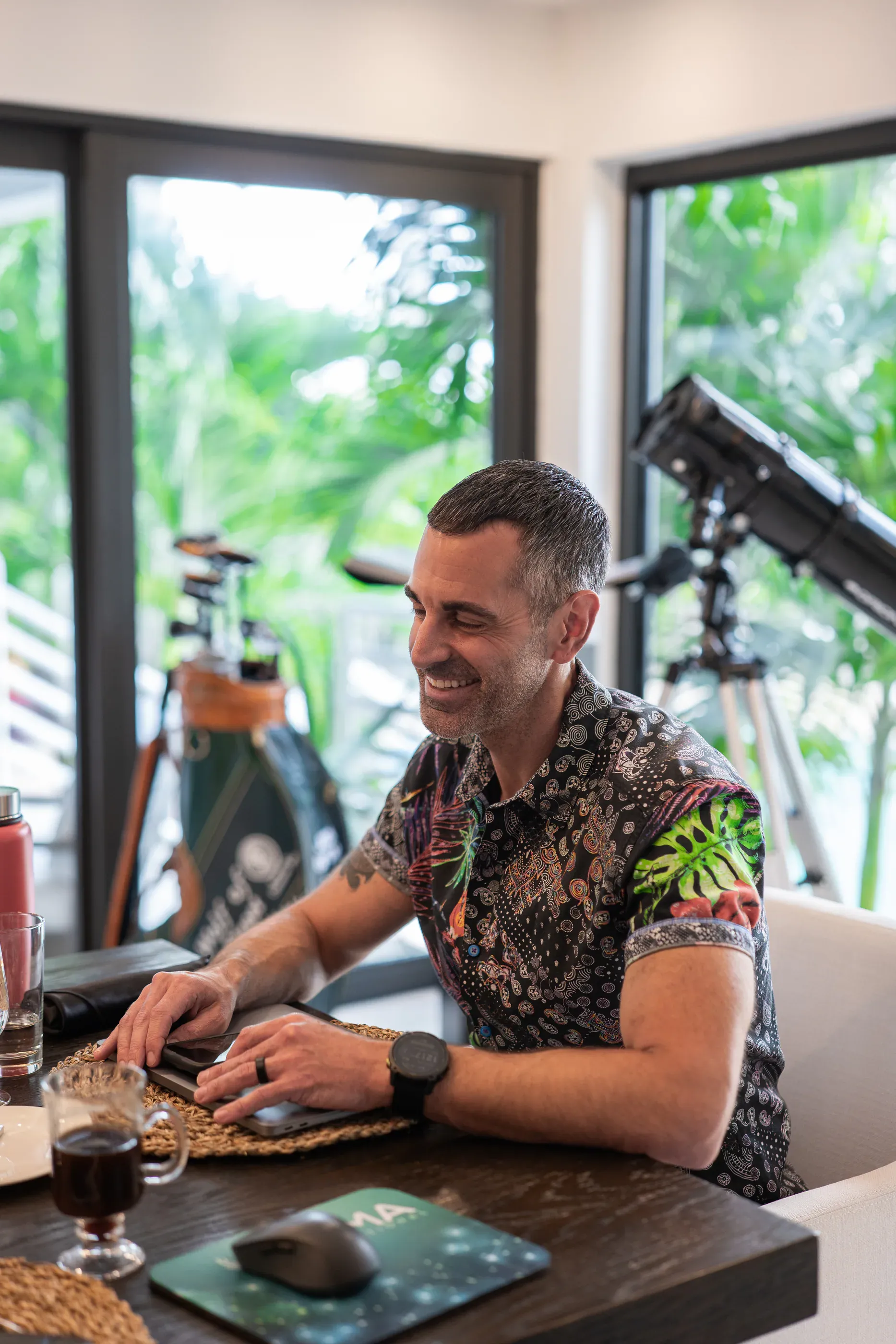 A guest smiling at his laptop at the dining table, with tropical greenery through the windows behind him.