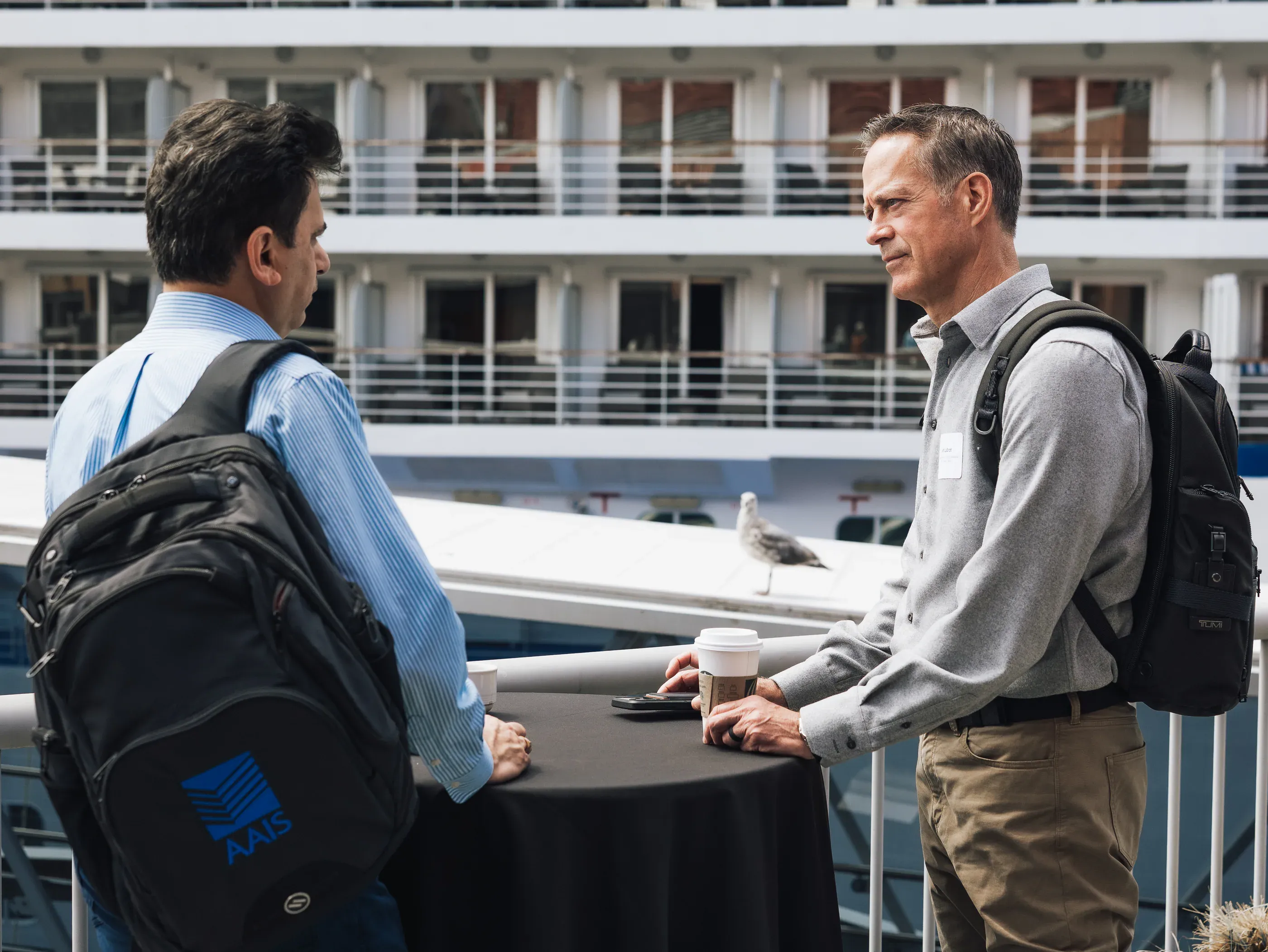 Two attendees talking at a cocktail table on a waterfront terrace with a cruise ship behind them.