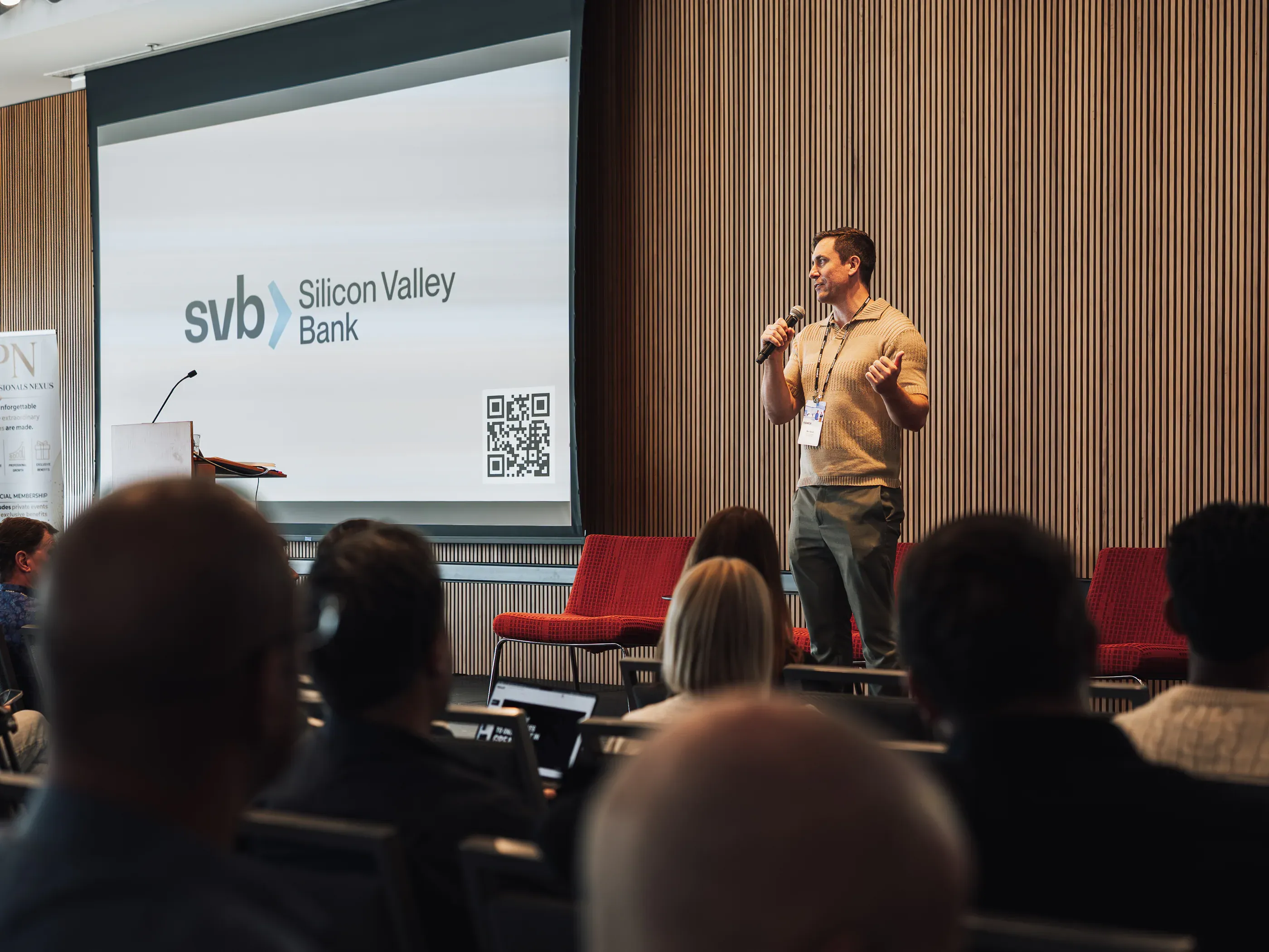 Speaker with a microphone on stage in front of a sponsor slide.